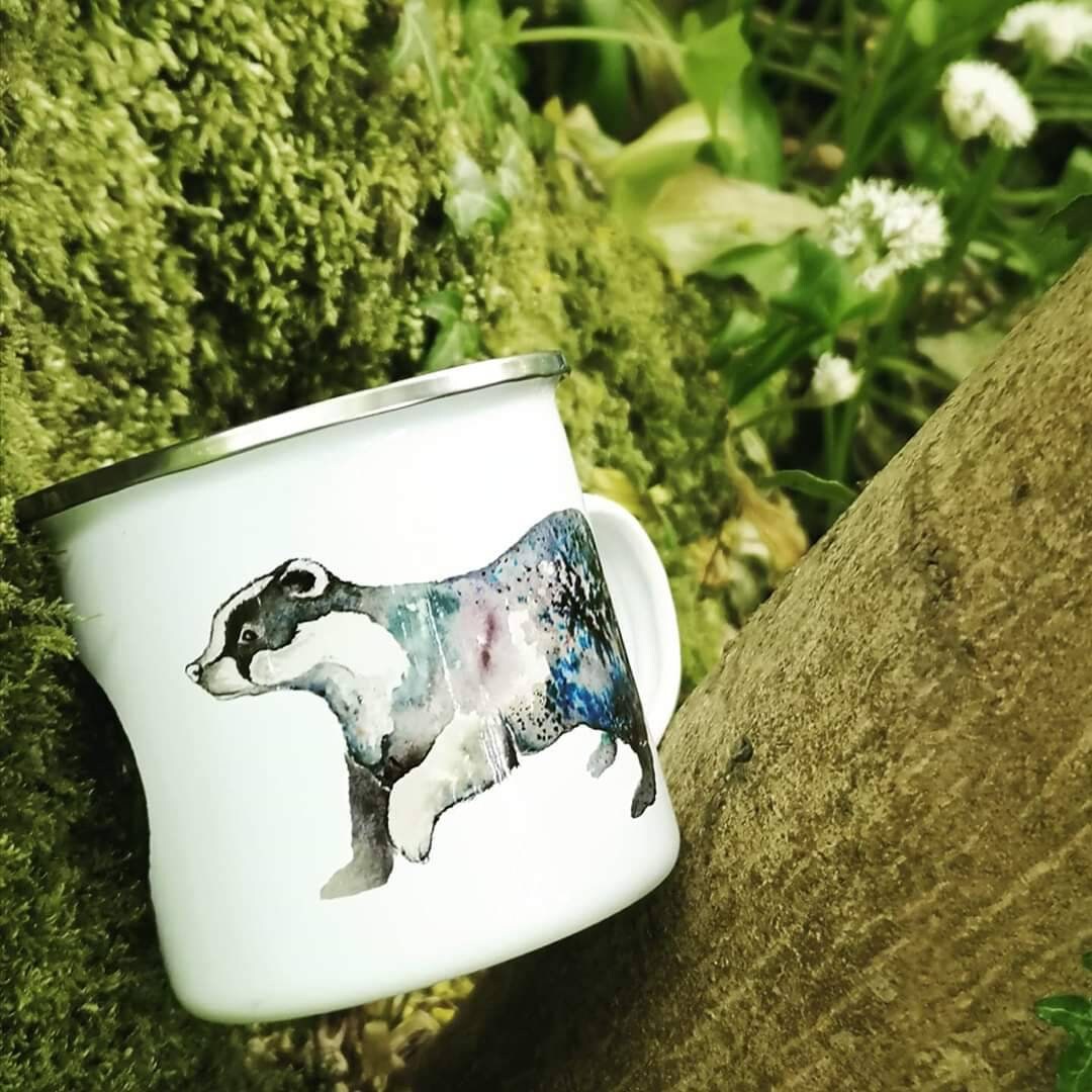 Enamel mug with a badger illustration resting on some moss with woodland flowers behind. A great mug you can take on a walk.