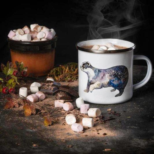 An enamel mug with a badger illustration from an original watercolour painting sits in the foreground. The mug is filled with hot chocolate and has chocolate and marshmallows around it.