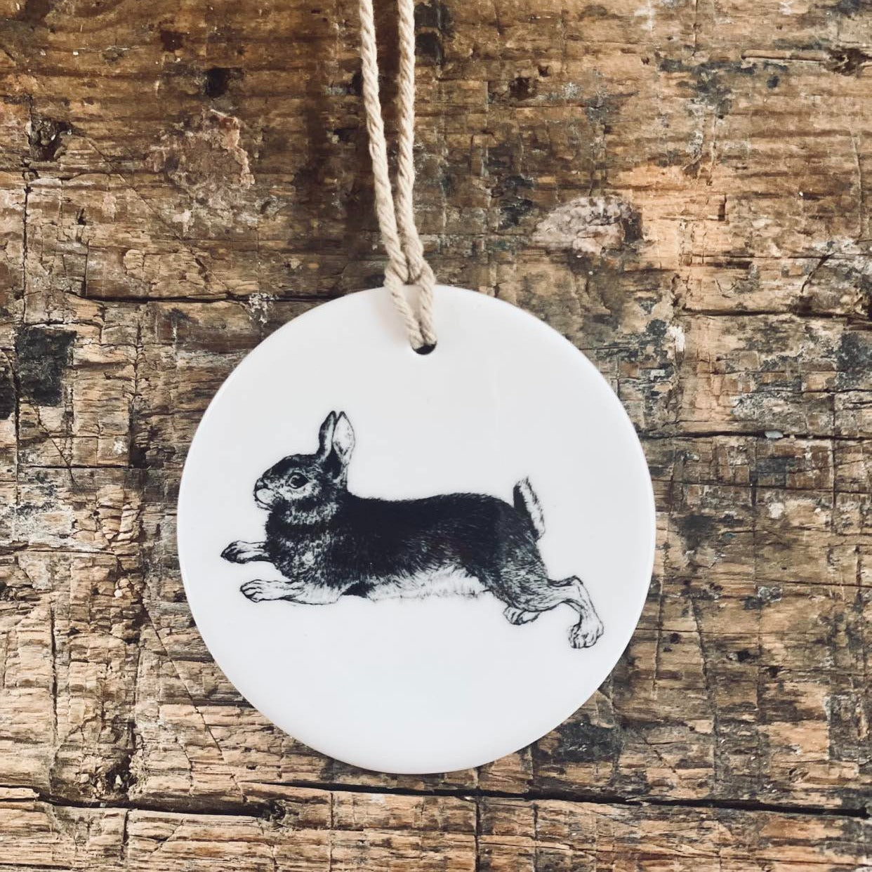 Round, while ceramic decoration with a handdrawn dwarf rabbit printed on in black. The bauble has a natural jute twine to hang it from and is placed on a rustic farmhouse table.