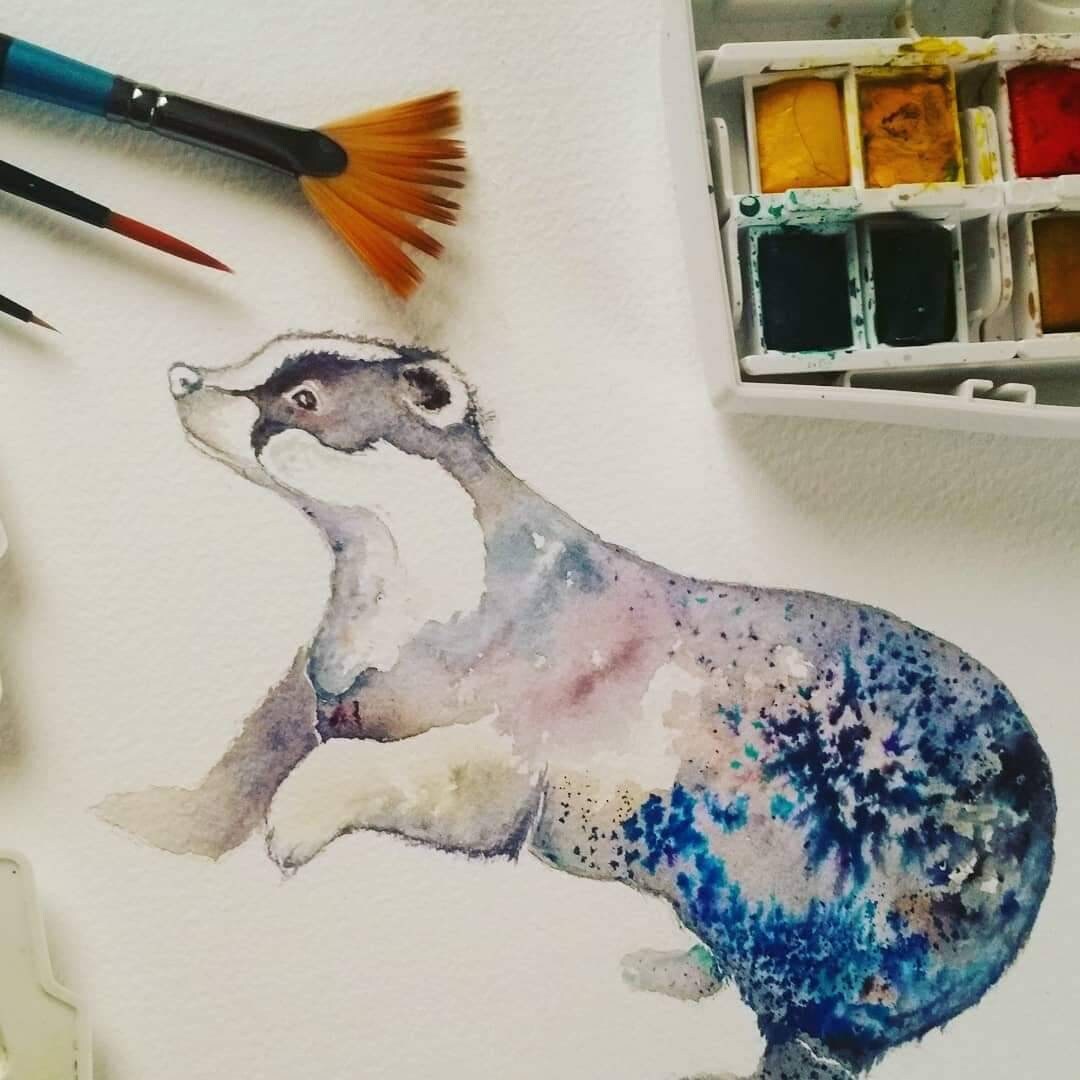 Original badger watercolour illustration with paints and brushes surrounding it.